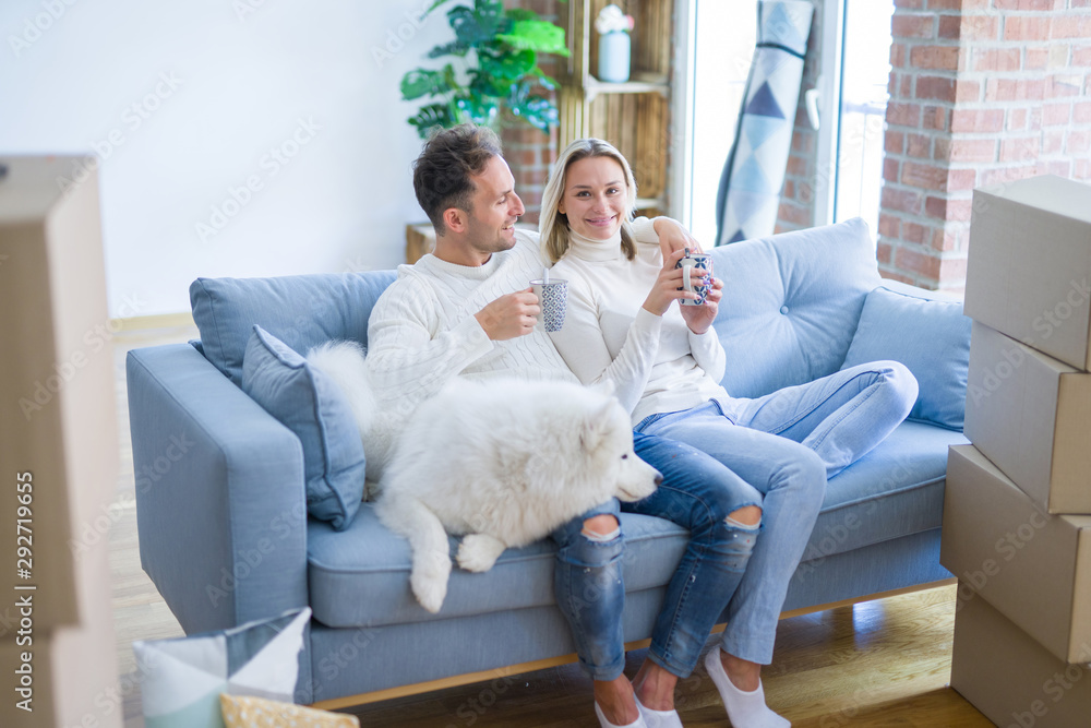 Young beautiful couple with dog sitting on the sofa drinking coffee at new home around cardboard boxes Young beautiful couple with dog sitting on the sofa drinking coffee at new home around cardboard boxes