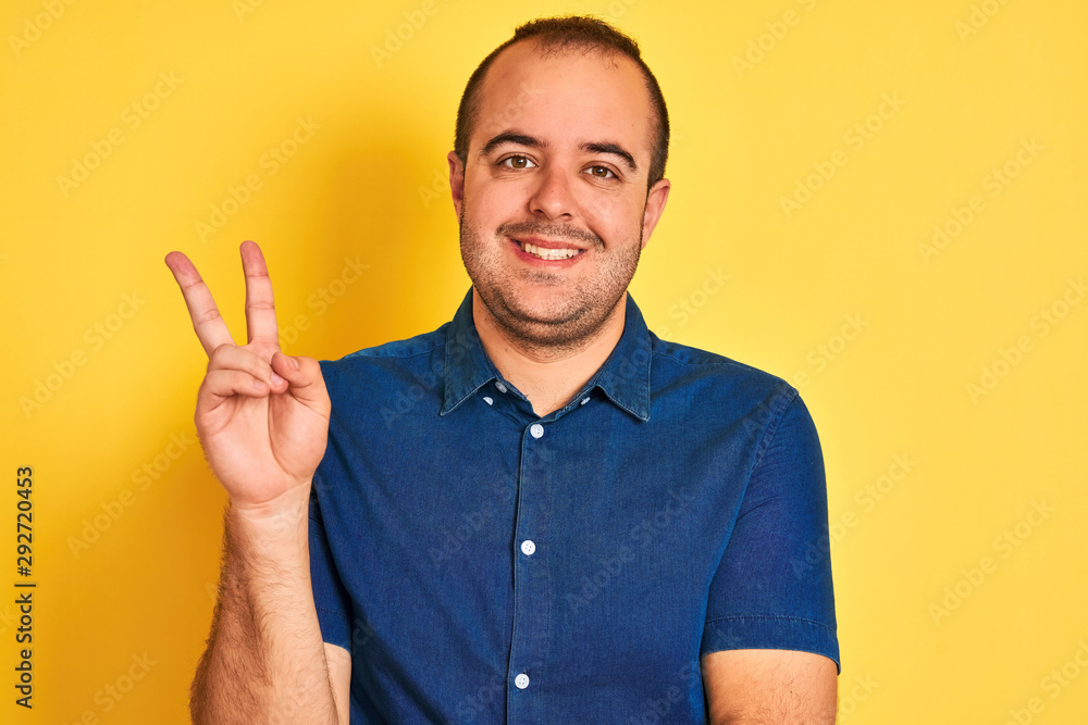 Young man wearing denim casual shirt standing over isolated yellow background smiling with happy face winking at the camera doing victory sign. Number two. Young man wearing denim casual shirt standing over isolated yellow background smiling with happy face winking at the camera doing victory sign. Number two.