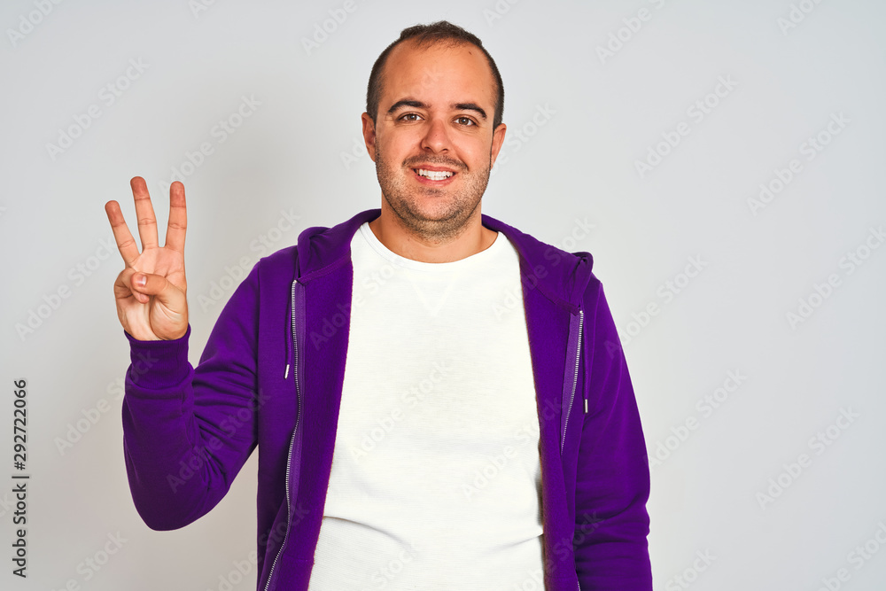 Young man wearing purple sweatshirt standing over isolated white background showing and pointing up with fingers number three while smiling confident and happy. Young man wearing purple sweatshirt standing over isolated white background showing and pointing up with fingers number three while smiling confident and happy.