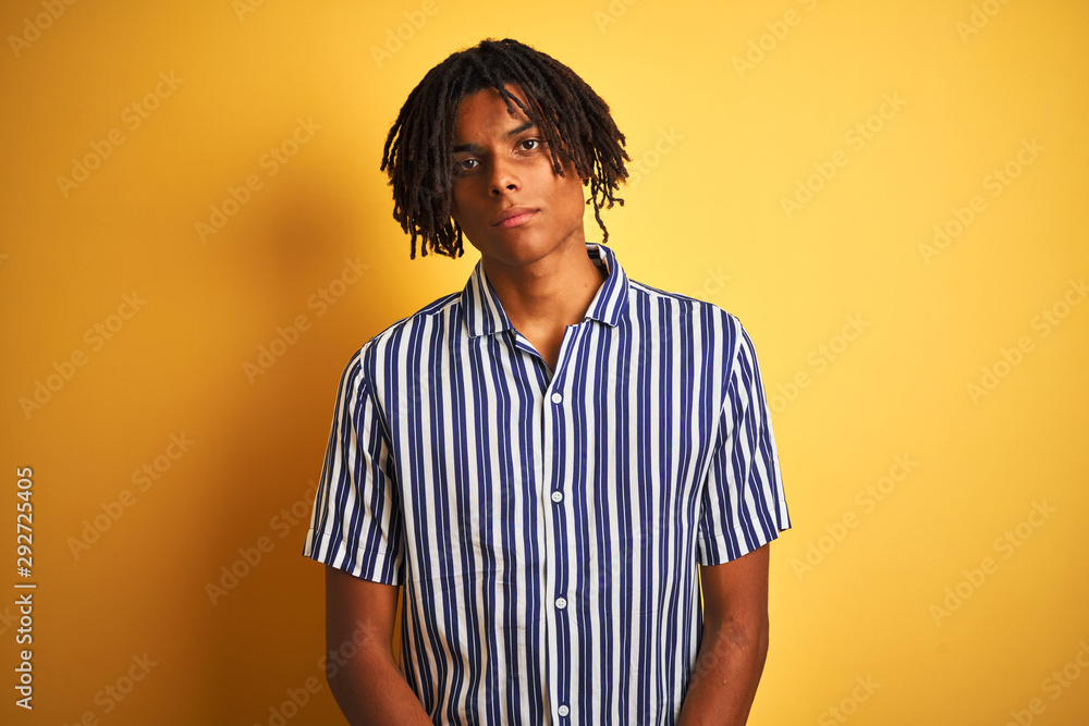 Afro man with dreadlocks wearing casual striped t-shirt over isolated yellow background Relaxed with serious expression on face. Simple and natural looking at the camera. Afro man with dreadlocks wearing casual striped t-shirt over isolated yellow background Relaxed with serious expression on face. Simple and natural looking at the camera.