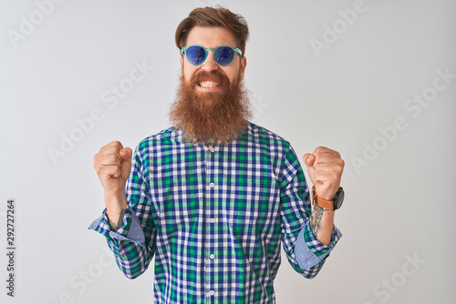 Young redhead irish man wearing casual shirt and sunglasses over isolated white background excited for success with arms raised and eyes closed celebrating victory smiling. Winner concept.