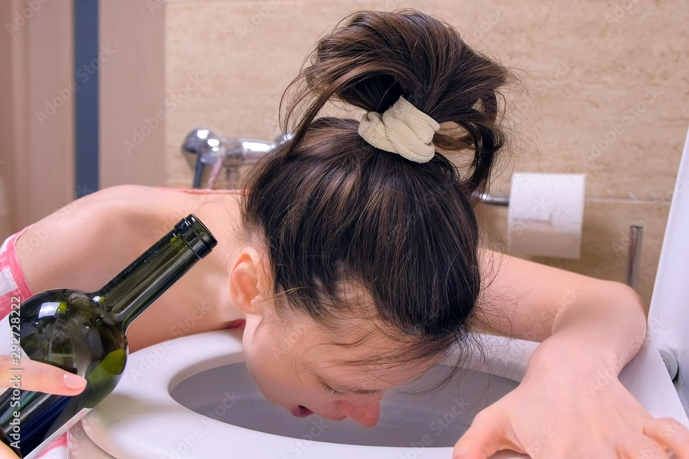Naklejka premium Drunk young woman is vomiting in toilet sitting on the floor with wine bottle in hand at home, morning hangover, side view.