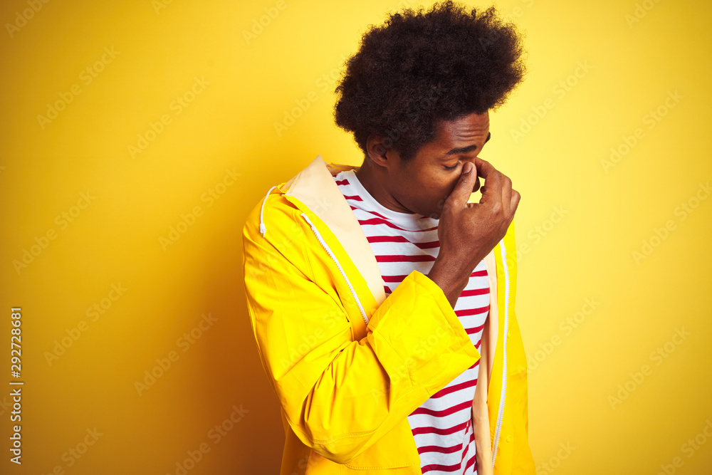 African american man with afro hair wearing rain coat standing over isolated yellow background tired rubbing nose and eyes feeling fatigue and headache. Stress and frustration concept. African american man with afro hair wearing rain coat standing over isolated yellow background tired rubbing nose and eyes feeling fatigue and headache. Stress and frustration concept.
