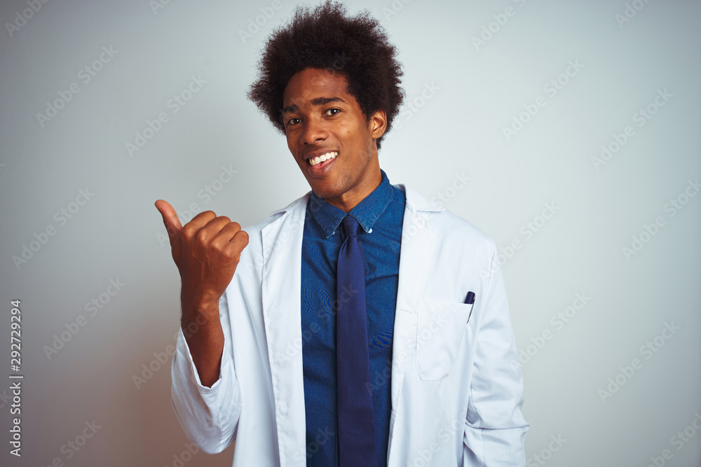 Young african american doctor man wearing coat standing over isolated white background smiling with happy face looking and pointing to the side with thumb up. Young african american doctor man wearing coat standing over isolated white background smiling with happy face looking and pointing to the side with thumb up.