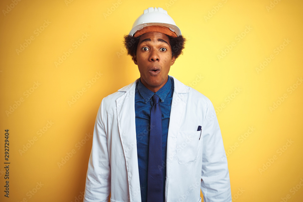 Afro american engineer man wearing white coat and helmet over isolated yellow background afraid and shocked with surprise expression, fear and excited face. Afro american engineer man wearing white coat and helmet over isolated yellow background afraid and shocked with surprise expression, fear and excited face.
