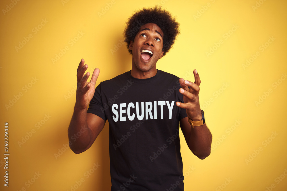 American safeguard man with afro hair wearing security uniform over isolated yellow background crazy and mad shouting and yelling with aggressive expression and arms raised. Frustration concept. American safeguard man with afro hair wearing security uniform over isolated yellow background crazy and mad shouting and yelling with aggressive expression and arms raised. Frustration concept.
