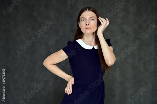 Portrait of a pretty young brunette woman with combed hair on a gray background in a dark blue dress. It is in different poses. Looks straight at the camera. Slim figure.