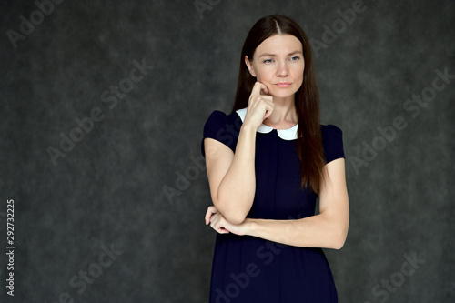 Portrait of a pretty young brunette woman with combed hair on a gray background in a dark blue dress. It is in different poses. Looks straight at the camera. Slim figure.