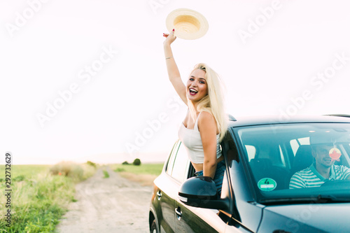 Happy woman traveling by car. Road trip travel vacation. Young woman leaning out car window with straw hat in hand.