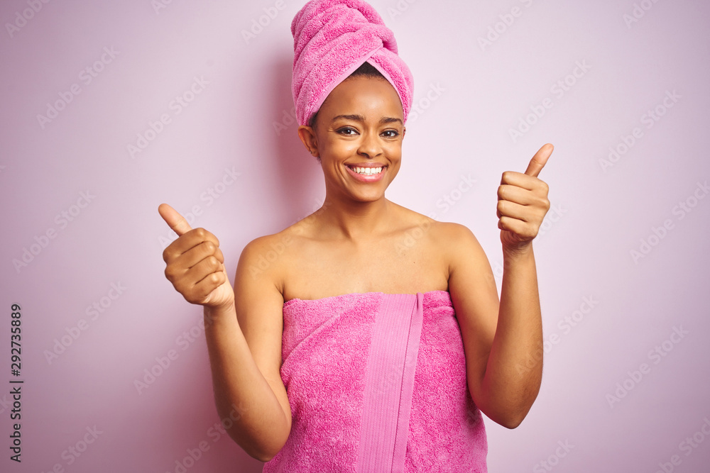African american woman wearing shower towel after bath over pink isolated background success sign doing positive gesture with hand, thumbs up smiling and happy. Cheerful expression and winner gesture. African american woman wearing shower towel after bath over pink isolated background success sign doing positive gesture with hand, thumbs up smiling and happy. Cheerful expression and winner gesture.