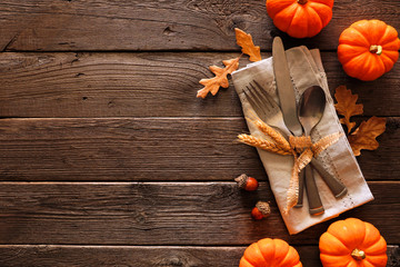 Autumn harvest or thanksgiving table scene with silverware, napkin, leaves and pumpkin border against a rustic dark wood background