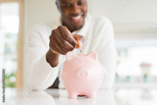 Handsome young african man putting a coin inside piggy bank smiling, excited about investment photo