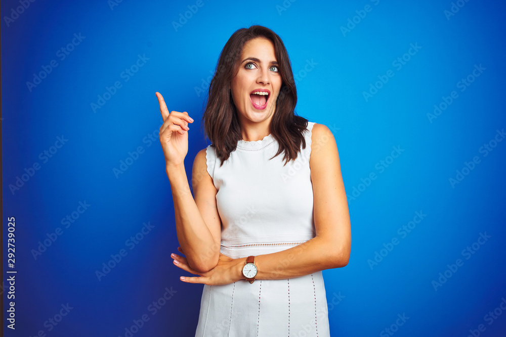 Young business woman wearing white elegant dress standing over blue isolated background with a big smile on face, pointing with hand and finger to the side looking at the camera. Young business woman wearing white elegant dress standing over blue isolated background with a big smile on face, pointing with hand and finger to the side looking at the camera.