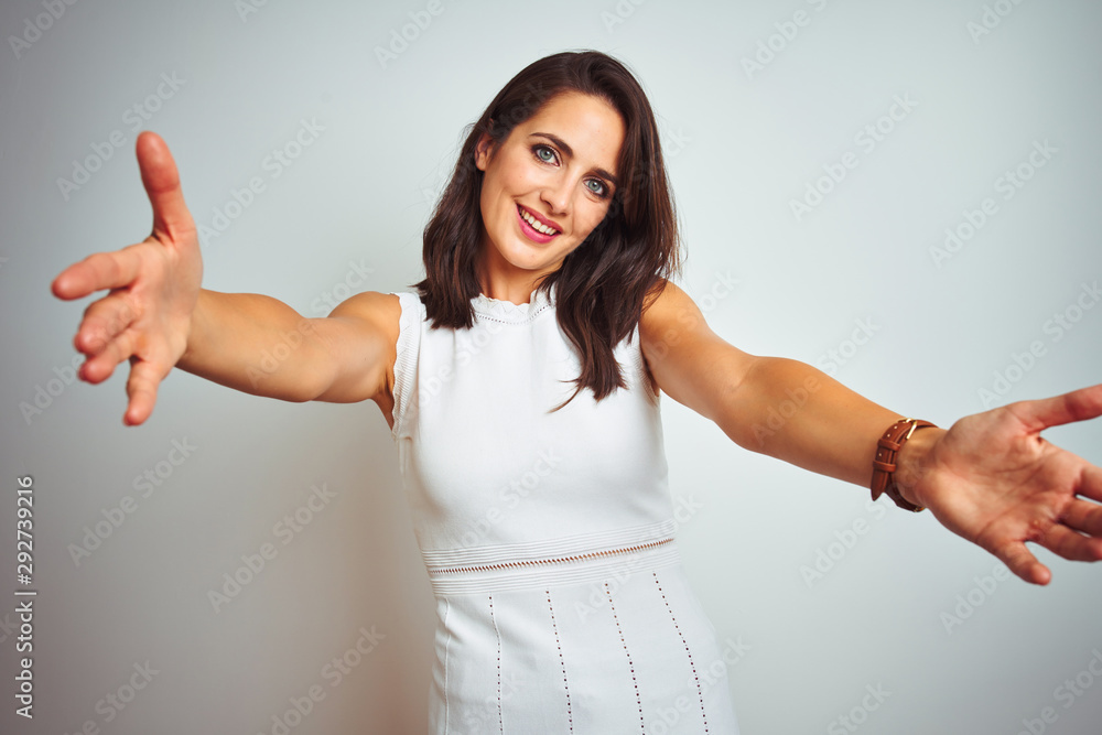 Young beautiful woman wearing dress standing over white isolated background looking at the camera smiling with open arms for hug. Cheerful expression embracing happiness. Young beautiful woman wearing dress standing over white isolated background looking at the camera smiling with open arms for hug. Cheerful expression embracing happiness.