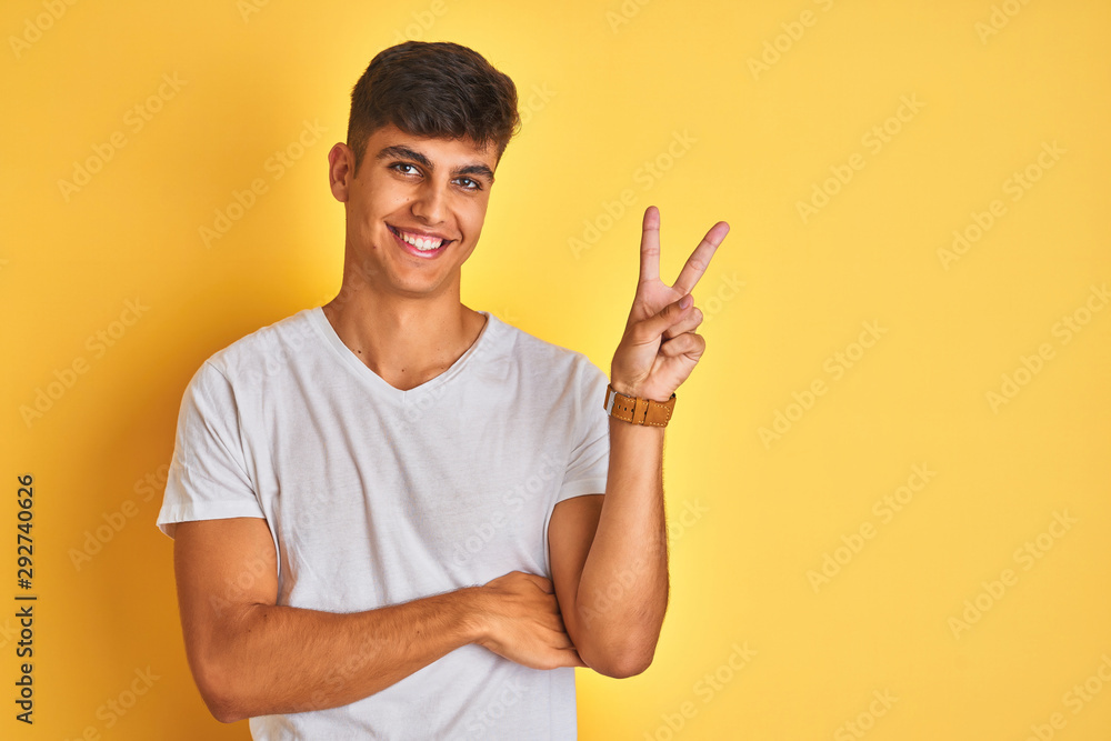 Young indian man wearing white t-shirt standing over isolated yellow background smiling with happy face winking at the camera doing victory sign with fingers. Number two. Young indian man wearing white t-shirt standing over isolated yellow background smiling with happy face winking at the camera doing victory sign with fingers. Number two.