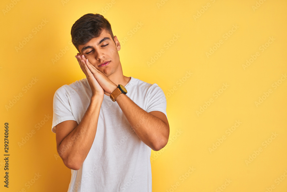 Young indian man wearing white t-shirt standing over isolated yellow background sleeping tired dreaming and posing with hands together while smiling with closed eyes. Young indian man wearing white t-shirt standing over isolated yellow background sleeping tired dreaming and posing with hands together while smiling with closed eyes.