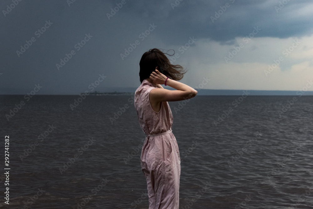 Fototapeta premium A cute young girl in a pink dress is standing in the water with flying hair during a storm and bad weather. Epic photo. Back view