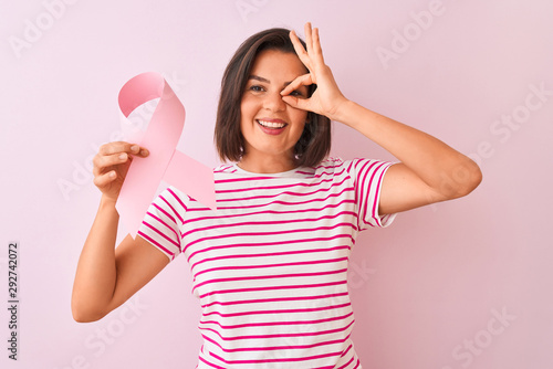 Young beautiful woman holding cancer ribbon standing over isolated pink background with happy face smiling doing ok sign with hand on eye looking through fingers