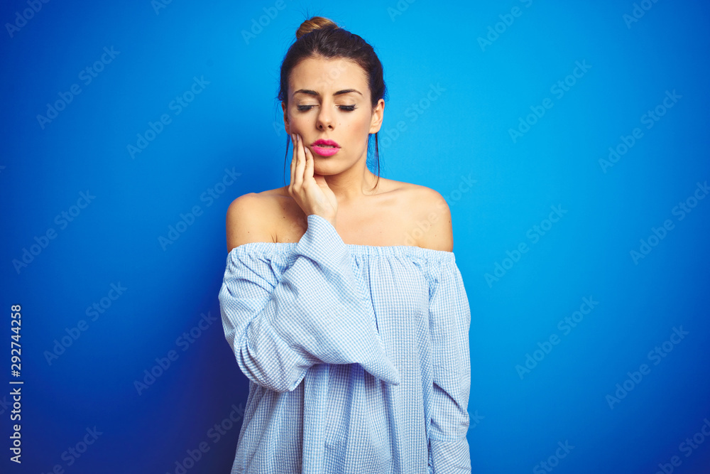 Young beautiful woman wearing bun hairstyle over blue isolated background touching mouth with hand with painful expression because of toothache or dental illness on teeth. Dentist concept. Young beautiful woman wearing bun hairstyle over blue isolated background touching mouth with hand with painful expression because of toothache or dental illness on teeth. Dentist concept.