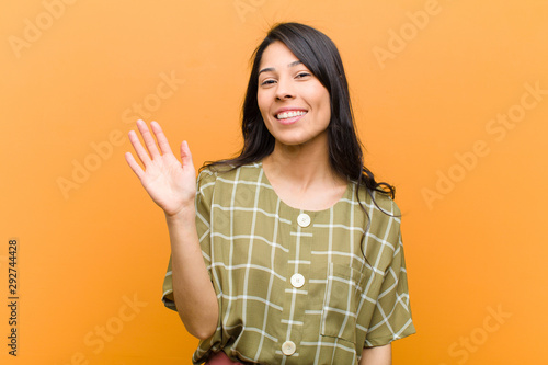 young pretty hispanic woman smiling happily and cheerfully, waving hand, welcoming and greeting you, or saying goodbye against brown wall photo