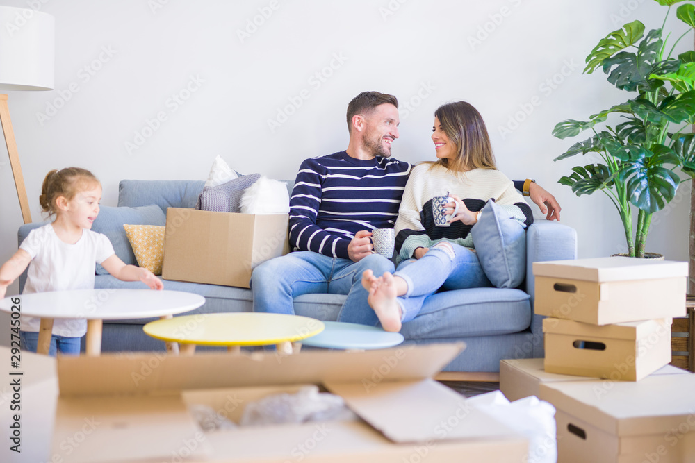 Beautiful family, parents sitting on the sofa drinking coffee looking his kid playing at new home around cardboard boxes Beautiful family, parents sitting on the sofa drinking coffee looking his kid playing at new home around cardboard boxes