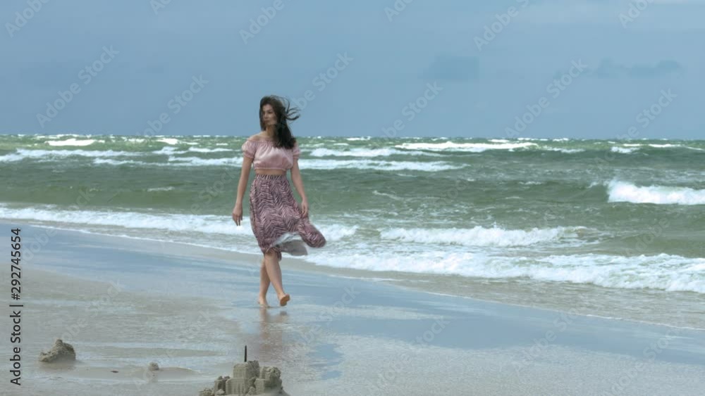 Tall brunette woman walking by the beach as the waves crashes. Slow motion Tall brunette woman walking by the beach as the waves crashes. Slow motion