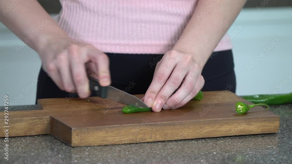 close up shot of female model's hand with kitchen knife cutting Green chili on a wooden board. she is wearing a pink top and black pants close up shot of female model's hand with kitchen knife cutting Green chili on a wooden board. she is wearing a pink top and black pants