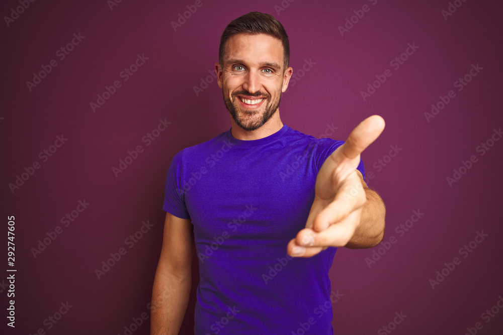 Young man wearing casual purple t-shirt over lilac isolated background smiling friendly offering handshake as greeting and welcoming. Successful business. Young man wearing casual purple t-shirt over lilac isolated background smiling friendly offering handshake as greeting and welcoming. Successful business.