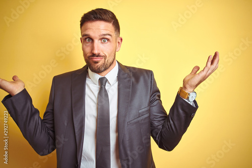 Young handsome business man over yellow isolated background clueless and confused expression with arms and hands raised. Doubt concept.