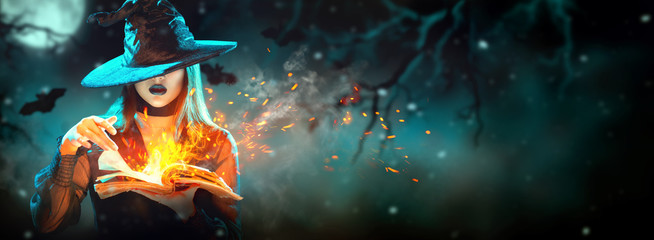 Halloween Witch girl with magic Book of spells portrait. Beautiful young woman in witches hat conjuring, making witchcraft. Over spooky dark magic forest background. Wide Halloween party art design.