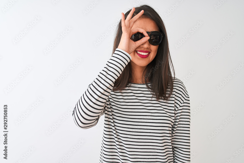 Chinese woman wearing striped t-shirt and sunglasses standing over isolated white background doing ok gesture with hand smiling, eye looking through fingers with happy face. Chinese woman wearing striped t-shirt and sunglasses standing over isolated white background doing ok gesture with hand smiling, eye looking through fingers with happy face.