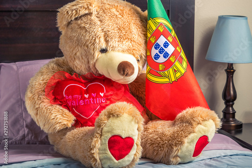 A Portugeuse teddy bear sitting on a bed with a heart that says 'Be my valentine'. photo