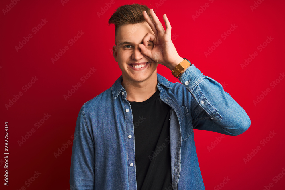Naklejka premium Young handsome man wearing denim shirt standing over isolated red background doing ok gesture with hand smiling, eye looking through fingers with happy face.
