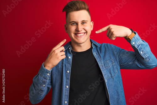 Young handsome man wearing denim shirt standing over isolated red background smiling cheerful showing and pointing with fingers teeth and mouth. Dental health concept.