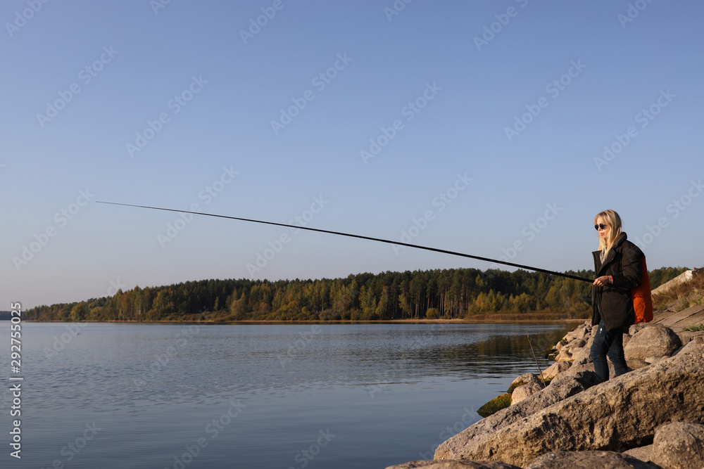 A beautiful blonde is fishing from the rocky shore of the lake in autumn