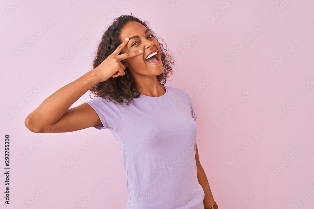 Young brazilian woman wearing t-shirt standing over isolated pink background Doing peace symbol with fingers over face, smiling cheerful showing victory Young brazilian woman wearing t-shirt standing over isolated pink background Doing peace symbol with fingers over face, smiling cheerful showing victory