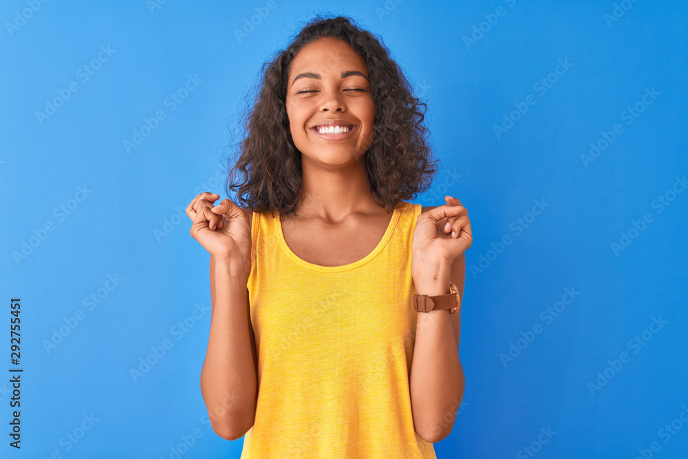 Young brazilian woman wearing yellow t-shirt standing over isolated blue background gesturing finger crossed smiling with hope and eyes closed. Luck and superstitious concept. Young brazilian woman wearing yellow t-shirt standing over isolated blue background gesturing finger crossed smiling with hope and eyes closed. Luck and superstitious concept.