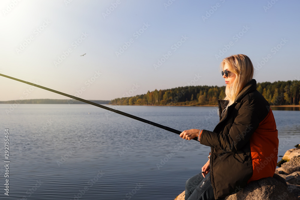 A beautiful blonde is fishing from the rocky shore of the lake in autumn A beautiful blonde is fishing from the rocky shore of the lake in autumn