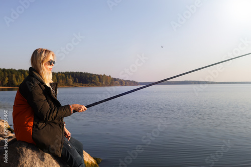 A beautiful blonde is fishing from the rocky shore of the lake in autumn
