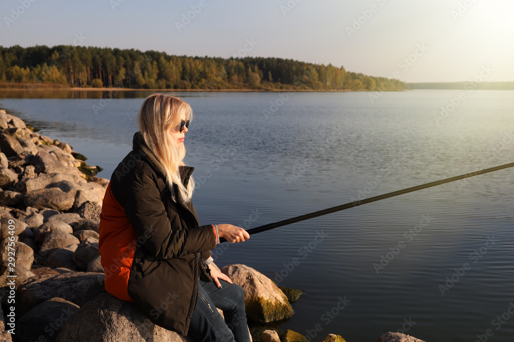 A beautiful blonde is fishing from the rocky shore of the lake in autumn A beautiful blonde is fishing from the rocky shore of the lake in autumn