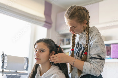 Girl braiding friend's hair in pigtail hairstyle sits on chair in room in front of mirror, window background photo