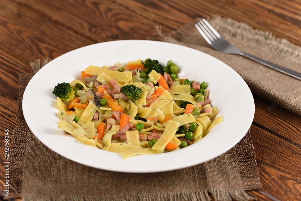 Noodle with broccoli, ham, carrots and peas in white dish.