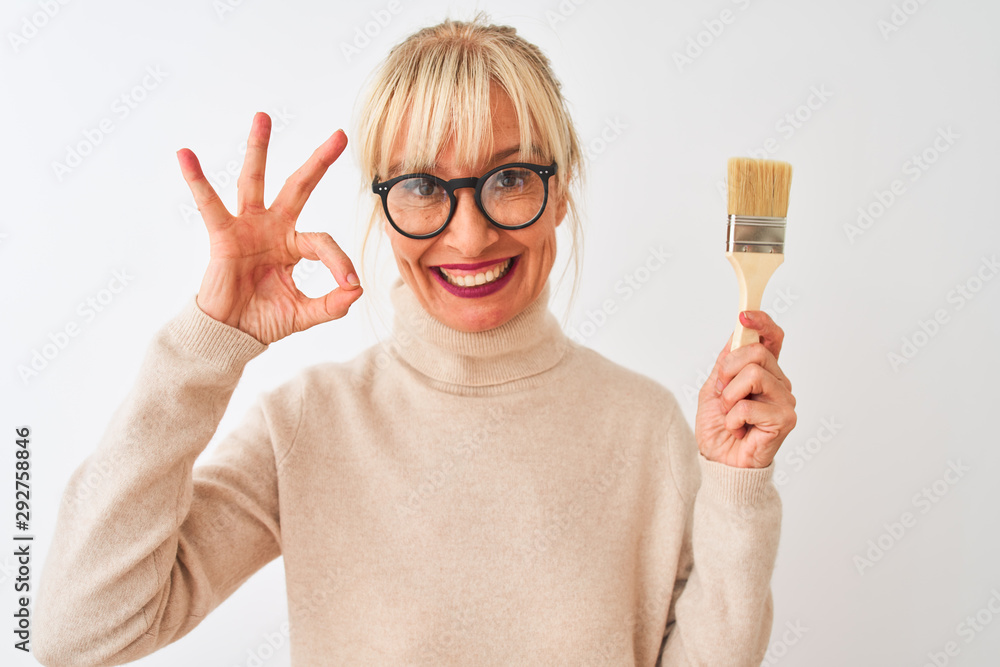 Middle age painter woman holding paint brush standing over isolated white background doing ok sign with fingers, excellent symbol Middle age painter woman holding paint brush standing over isolated white background doing ok sign with fingers, excellent symbol