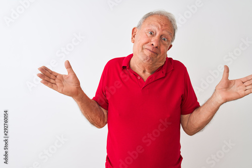 Senior grey-haired man wearing red polo standing over isolated white background clueless and confused expression with arms and hands raised. Doubt concept.