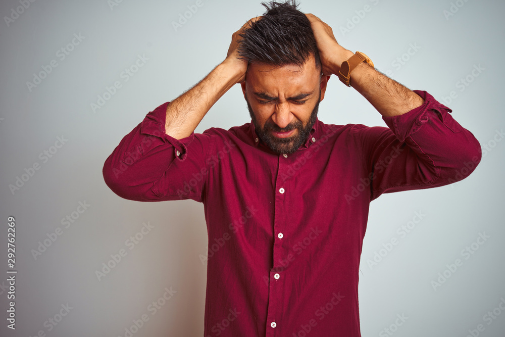Young indian man wearing red elegant shirt standing over isolated grey background suffering from headache desperate and stressed because pain and migraine. Hands on head. Young indian man wearing red elegant shirt standing over isolated grey background suffering from headache desperate and stressed because pain and migraine. Hands on head.