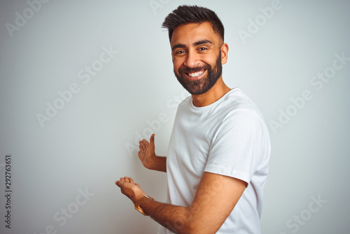 Young indian man wearing t-shirt standing over isolated white background Inviting to enter smiling natural with open hand photo