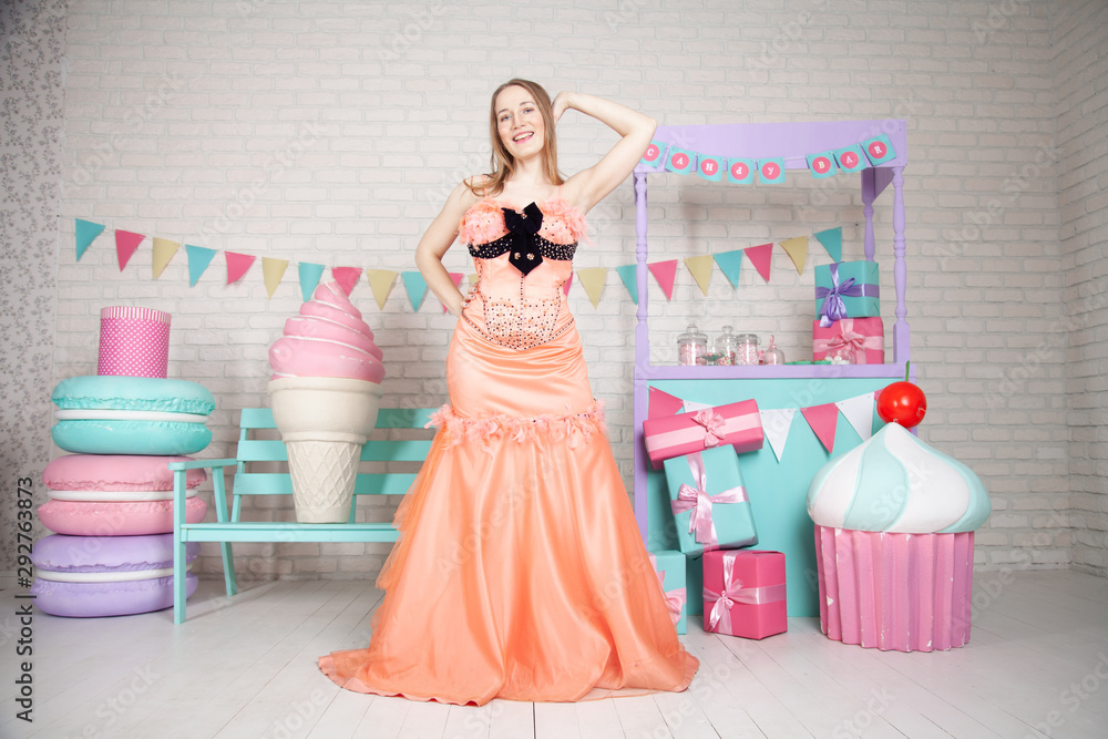cute happy adult girl posing in fashionable dress among huge fabulous sweets and cakes cute happy adult girl posing in fashionable dress among huge fabulous sweets and cakes