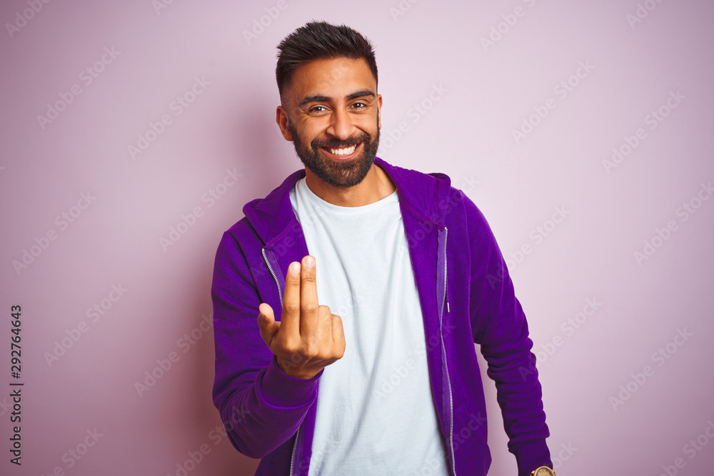 Young indian man wearing purple sweatshirt standing over isolated pink background Beckoning come here gesture with hand inviting welcoming happy and smiling - obrazy, fototapety, plakaty 
