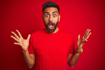 Young handsome indian man wearing t-shirt over isolated red background celebrating crazy and amazed for success with arms raised and open eyes screaming excited. Winner concept