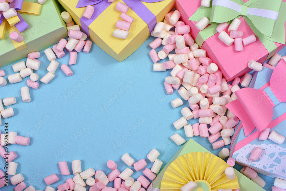 Flat lay background with pastel colored paper craft gift boxes with ribbons and marshmallowe sweets framing copy space in middle of blue background